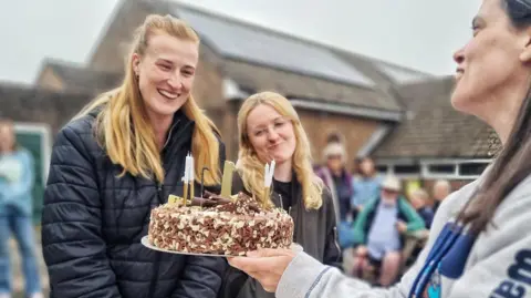 Handout Two blonde women, in black puffer jackets, looking at a chocolate cake which is held by a woman wearing a grey hoodie. There are people standing or sitting in the background.