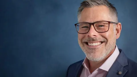 One to One Personnel A professional-looking headshot of man with a with glasses who is smiling and wearing a suit with a blue background