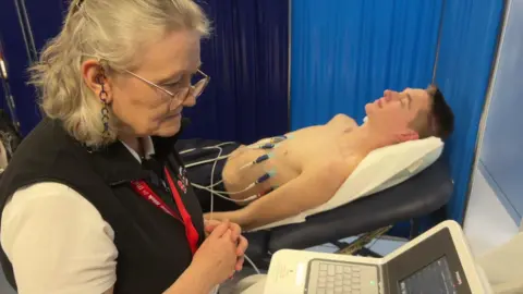 A woman with a red lanyard on looking at a computer and a boy on a medical bed behind her with wires attached to his chest.