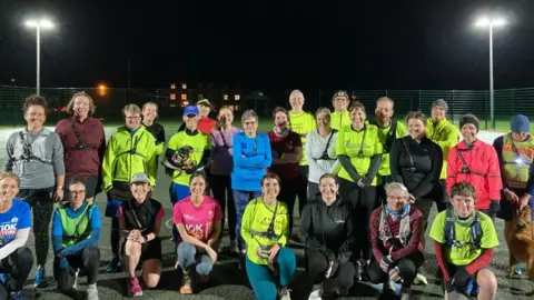 Haxby Runners pose for the camera - a mixed gendered group, wearing fluorescent running gear. In the background, a sports court can be seen, lit by large floodlights. 