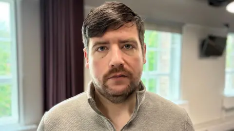 Alan Irwin had a beige half zip jumper on, He has a brown moustache and beard, and his hair is swept over to one side.