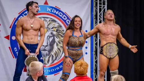 Nadia Sapphire Two wrestlers, both holding or wearing belts, accompanying a woman in the centre, wearing a leopard-print bikini-type outfit