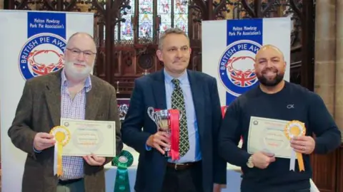 Baldy's Dark Kitchen Alex Melling from Baldy's Dark Kitchen stands holding a certificate to the right of a competition judge and another competition winner at the British Pie Awards. 