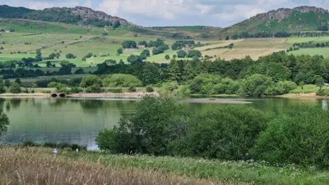 Severn Trent Water A reservoir viewed from a hill in the countryside. Across the water are a large number of trees and wooded areas with hills and fields stretching into the background.