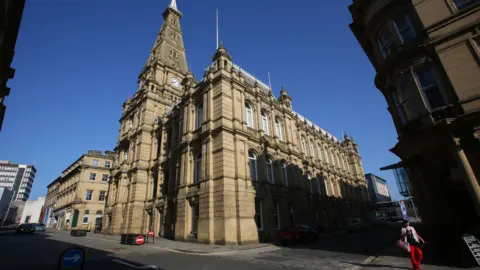 The outside of Halifax Town Hall - a tall beige stone building bathed in sunshine.