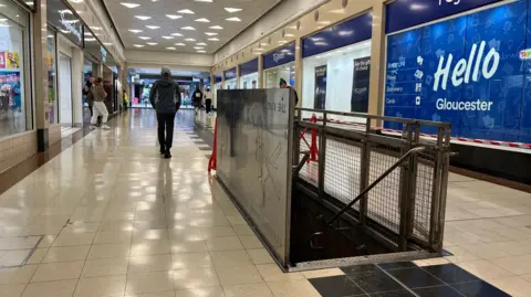 The inside of a shopping centre. There is a hole in the floor with stairs leading down it. There are people walking in the shopping centre.