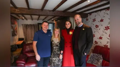 Princeandprincessofwales/Instagram (left to right) Butcher Kevin Kidner, Zsuzsanna, Catherine and William.
The four of them are stood in front of red sofas in a room with beams on the ceiling. Kevin is wearing a blue t-shirt and blue jeans, Zsuzsanna is wearing a leopard print dress under a black cardigan, Catherine is wearing a red coat with a daffodil pin on the left lapel, and William has blue trousers, a maroon jumper under a black blazer with a daffodil on the left lapel.