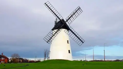 Little Marton Windmill, on a grassed area with a grey cloudy sky behind. The mill is white with four black sails and a black capped roof.