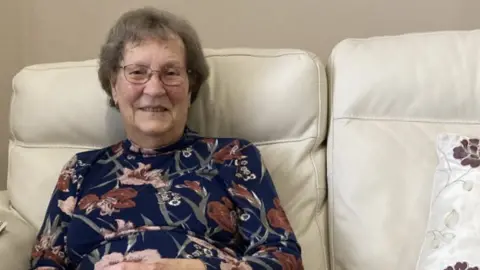 Barbara Quevâtre sitting on a cream leather sofa sharing memories of her late husband and the diving board at the bathing pools. She is wearing a flowery top and grey trousers and is smiling. 