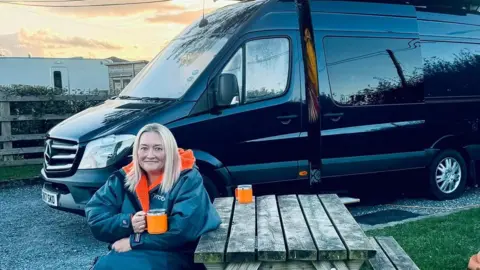Supplied Gayle Myers-Drake wearing a thick jacket sitting on a picnic bench while holding a mug containing a hot drink. Behind her is her large black campervan.