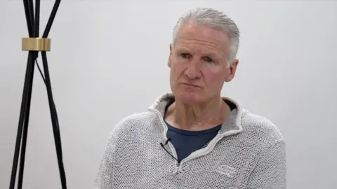 Deputy Tom Binet is wearing  a white cardigan with a blue T-shirt underneath. He is looking off camera and there is a black and gold lamp to his left.