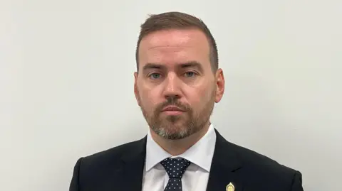 A head and shoulders picture of Basildon Borough Council leader Gavin Callaghan. He is looking at the camera and wearing a black suit jacket, a white shirt and a black and white tie