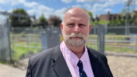A picture of the Mayor of Bedford Tom Wootton. He has a grey and white beard and moustache and is wearing a pink shirt and a black jacket and tie. He is stood in front of a railway line although the background is out of focus.