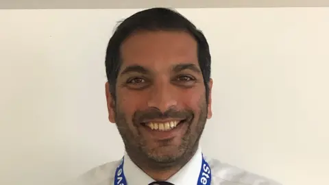 County Durham and Darlington NHS Foundation Trust Steve Russell, smiling brightly at the camera. He has dark, short hair and a light beard. He is wearing a white shirt and a blue lanyard.