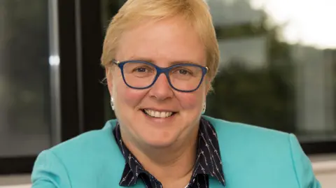 Worcestershire Acute Hospitals NHS Trust Portrait photograph of Dame Julie Moore wearing a light blue jacket and blue-framed glasses.