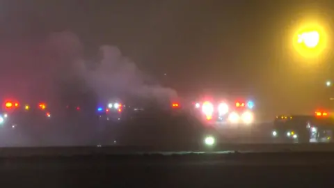The blurry image shows the runway at Bangor Airport with smoke rising and red lights emanating from vehicles nearby 