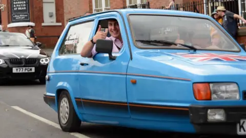 Harry Atkinson driving the powder blue Reliant Robin, smiling and giving a thumbs up out of the side window.