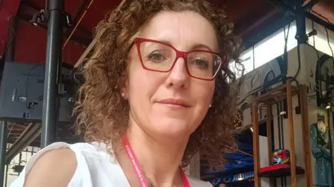 A woman with shoulder-length curly brown hair and red framed glasses looks into the camera.