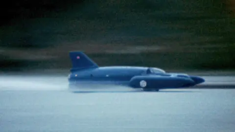 Neil Sheppard A close-up of Bluebird - a craft that looks like a cross between a plane and a boat. It is painted blue and is running at speed on Coniston Water. It appears to just skim the surface of the water.
