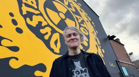 Jonn Penney has grey hair and is smiling at the camera. He wears a black shirt and T-shirt with writing on. Behind him is a yellow and black mural. 