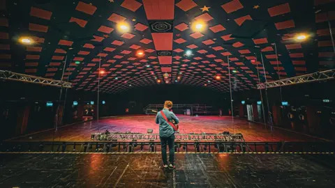 Andrew Dickson A man playing a guitar standing on the middle of a stage looking on to an empty auditorium