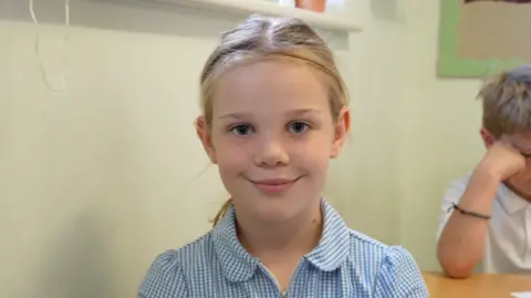 Qays Najm/BBC A young girl wearing a blue and white chequered school dress. She has blonde hair that has been tied back and she is smiling at the camera.