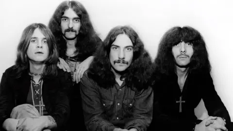 A black and white photograph of the band Black Sabbath - four men with long hair with three sitting in a line and one just behind them.