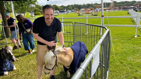 A man in a dark blue t-shirt and light brown shorts is holding the reins of a miniature pony within a gated area of the Three Counties Show. A fair is in the distance, with a show arena in front if that