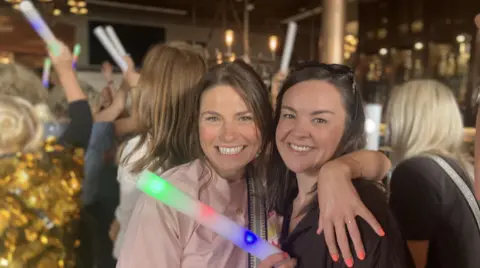 Two women smile with their arms around each other's shoulders. One holds a glow stick and they are dancing in the middle of a crowd.