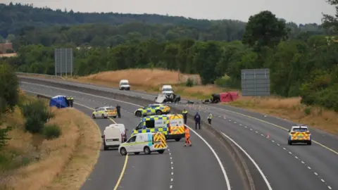 A wide shot of a car crash site. Many emergency service vehicles are on the road and debris from the crash.