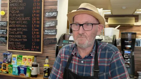 Frank Newson stands behind a cafe counter looking slightly away from the camera. He wears a straw bowler hat, glasses, a blue and red chequered shirt and a black apron. He has a grey beard. Behind him coffee machines and other cafe items can be seen.