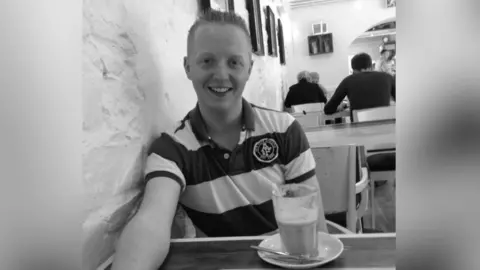 BELL FAMILY A black and white photo of Kevin Bell smiling at the camera in a cafe. He is sitting at a table in front of a half-full glass of a milky drink. He has short hair and is wearing a striped, short-sleeved polo shirt. There are other customers in the background. 