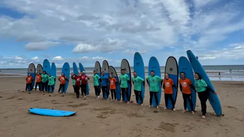Adela Rokytova A group of 17 people standing on the wet sand of the beach. Each has a surfboard that they are holding stood up behind their backs. The surfboards are blue and white. The majority of people are wearing either a turquoise or orange t-shirt over a wet-suit, and there are two people stood central who are wearing a red top.