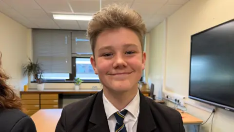 Etienne pictured with light brunette hair, short on the sides and long on the top. He is smiling with a closed mouth at the camera. Etienne is wearing a white shirt with a black blazer and navy striped tie.
