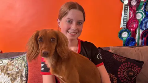 Taylor with long ginger hair tied back smiling at the camera and wearing a dark blue T-shirt. A brown cocker spaniel is sitting on her lap and looking at the camera. There are cushions behind them and an orange wall. There are competition rosettes in the top right of the picture.
