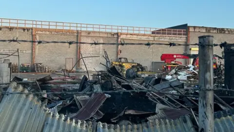 A large pile of fire-damaged scrap metal in front of a large wall. There is a yellow digger and a red fire truck in the background.