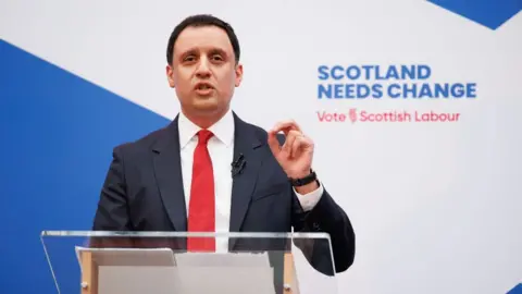 PA Media Anas Sarwar, who has short black hair, speaks at a podium while wearing a business suit and red tie. There is Labour branding behind him. 