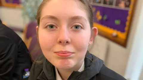 Amy has brown hair which is pulled back from her face. She is wearing a black blazer with a hooded top underneath.