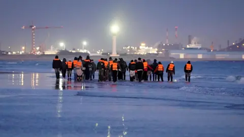 A group of around 20 people wearing lifejackets walking along the water's edge. 
