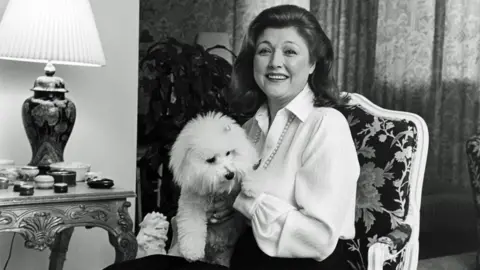 Getty Images Black-and-white photo of Barbara Taylor Bradford sitting in an ornate chair, smiling with a white poodle on her lap