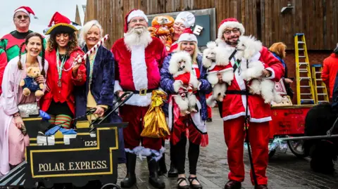 The Valley Evesham A group of people dressed in festive costumes. To the left is a cart which says "Polar Express" and a father Christmas is in the centre. To the right a man and woman in Christmas dress are holding three poodles. 