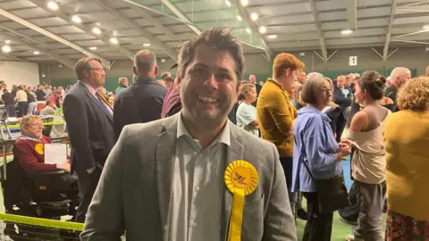 Leigh Frost wearing a grey blazer with a white striped shirt. He also has a yellow rosette on his left side with the Liberal Democrat bird logo on it. He has short brown hair and is stood in front of a crowd of people at the count.