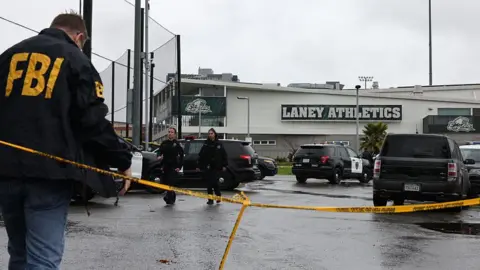A man in an FBI jacket stands near yellow police tape. A building in the background has a sign that says "Laney Athletics"