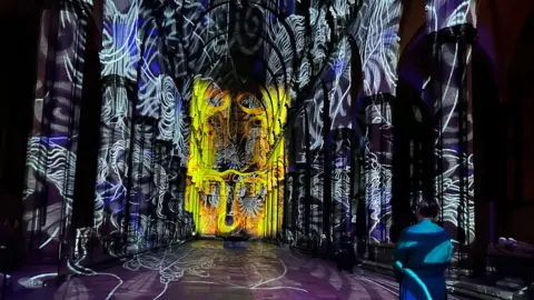 The dark inside of Salisbury Cathedral, a member of the clergy standing slightly to the side in shadow, but coloured swirling lights projected in blue all around the nave and bright yellow ones right at the front.