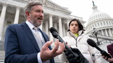 Thomas Massie speaks to the press with microphones and a reporter to his right in front of the U.S Capitol
