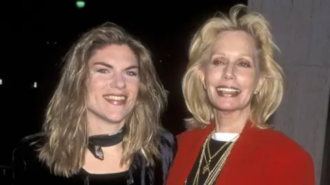 Claire Kellerman pictured with her mum actress Sally Kellerman in 1997. Claire has long, fair hair and wears a dark top and neck scarf. Sally has shoulder-length blonde hair and wears a black top, several gold necklaces and a red blazer. Both smile for the camera.