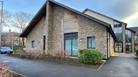 Lanchester Medical Centre is a beige brick building with a large triangle roof. It has large windows with Lanchester Medical Centre written across it with grey metal letters.