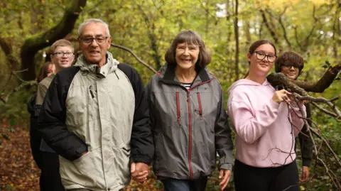 An older couple wearing cagoules walk hand in hand in the woods surrounded by teenagers. 