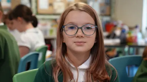 A school girl with long brown hair and blue rimmed glasses looks into the camera 
