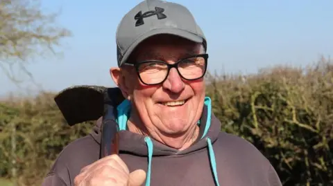 BBC An older man stood on Aldercar Lane, smiling for the camera in sunshine, with a shovel over his shoulders.